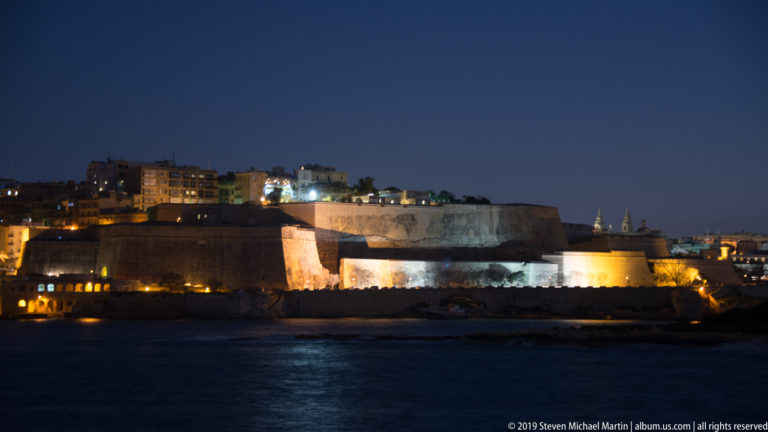 Floodlit limestone bastions of Fort Manoel across dark harbor water at night, with urban buildings rising behind.
