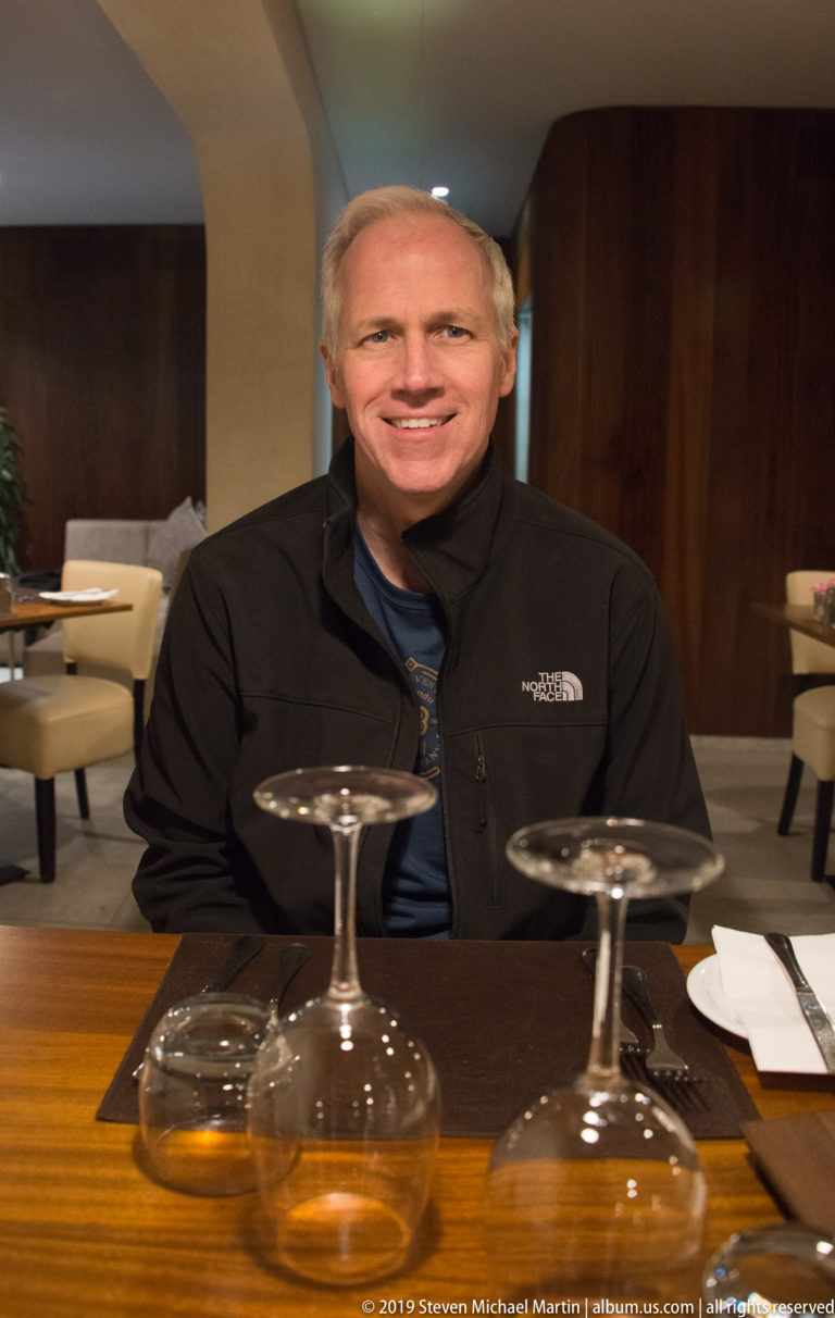 Smiling man in a dark jacket seated behind two upside-down stemmed glasses on a wooden restaurant table.