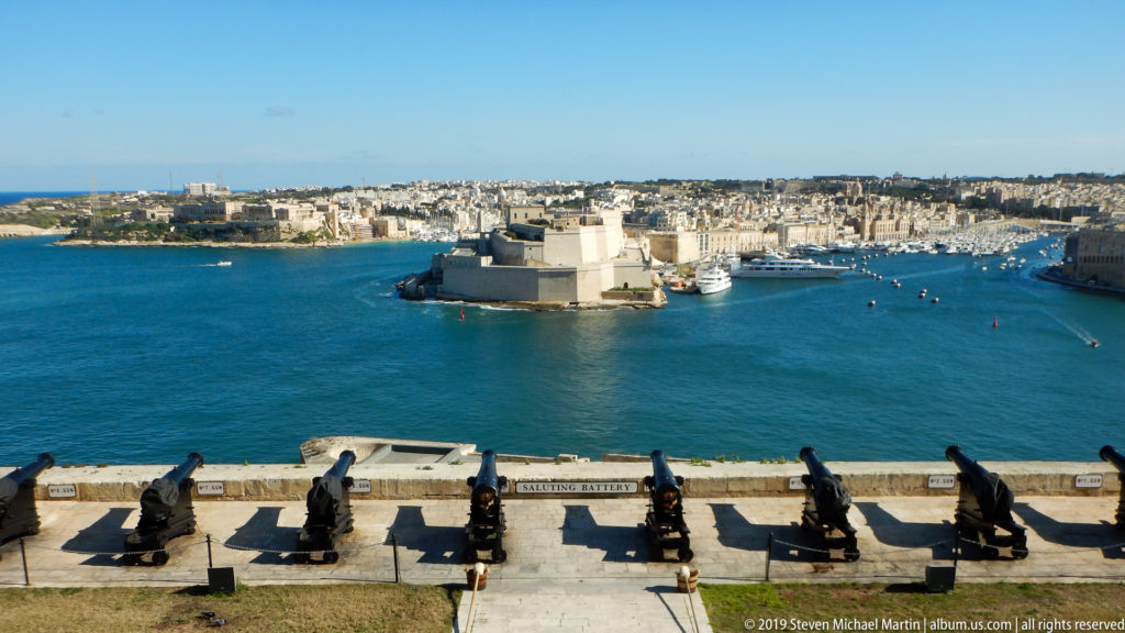 Row of historic cannon overlooking Grand Harbour, with Fort St Angelo centered amid Vittoriosa and marina-filled waterways.
