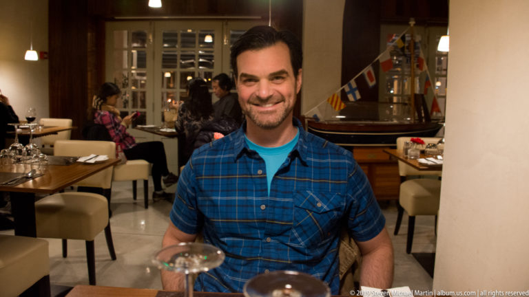 Smiling man in a blue plaid shirt seated at a restaurant table, with dining tables and small maritime flags behind him.