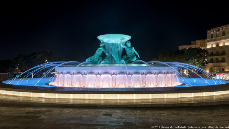 Full nighttime view of the Triton Fountain, with bronze figures, a glowing basin, and blue-lit water jets arcing around it.