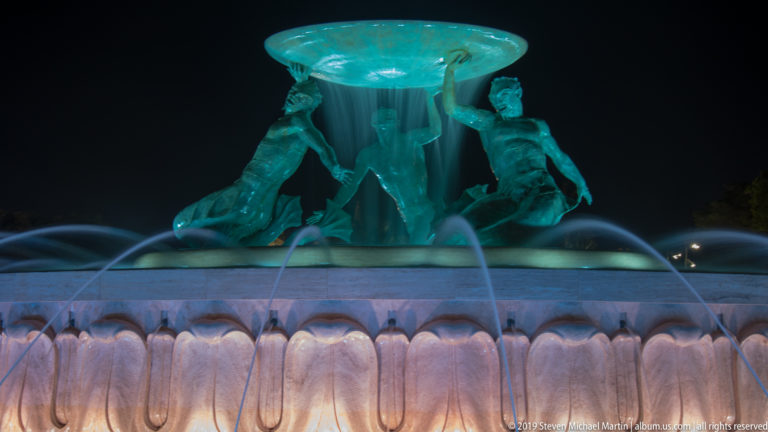 Illuminated bronze Triton figures support a large fountain basin as turquoise water and arcing jets glow against the night sky.