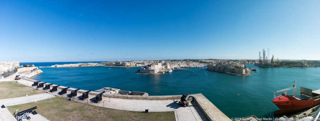 Elevated view across Grand Harbour from Valletta, with stone fortifications, artillery, blue water, and dense harbor-side development.