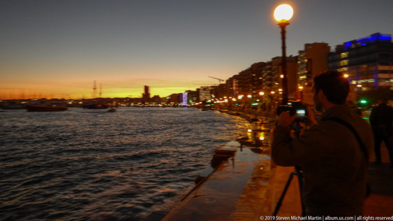 Person photographing a harbor sunset from a stone promenade lined with glowing streetlamps and waterfront buildings.