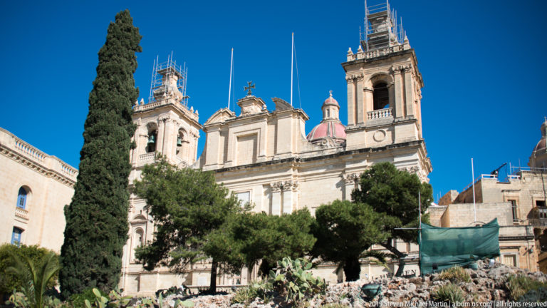 Ornate limestone church with twin bell towers, central dome, trees in front, and scaffolding on the right tower.