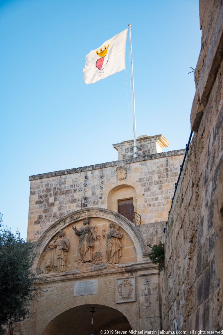 Maltese flag above a stone gateway with an arched relief of carved figures, framed by massive limestone walls.