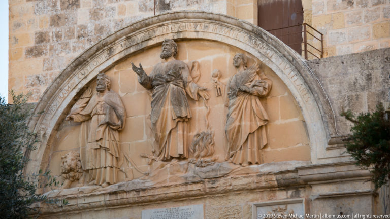 Arched limestone relief with three standing religious figures, a small central flame motif, and an inscription around the border.