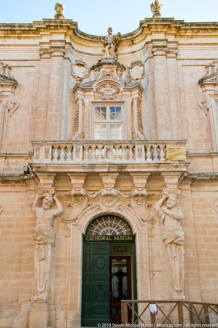 Ornately carved limestone entrance marked Cathedral Museum, with green doors, sculpted figures, balcony, balustrade, and curving Baroque details.