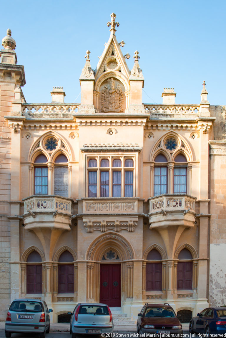 Symmetrical limestone palazzo with arched windows, carved balconies, red central door, steep ornamental gable, and parked cars.