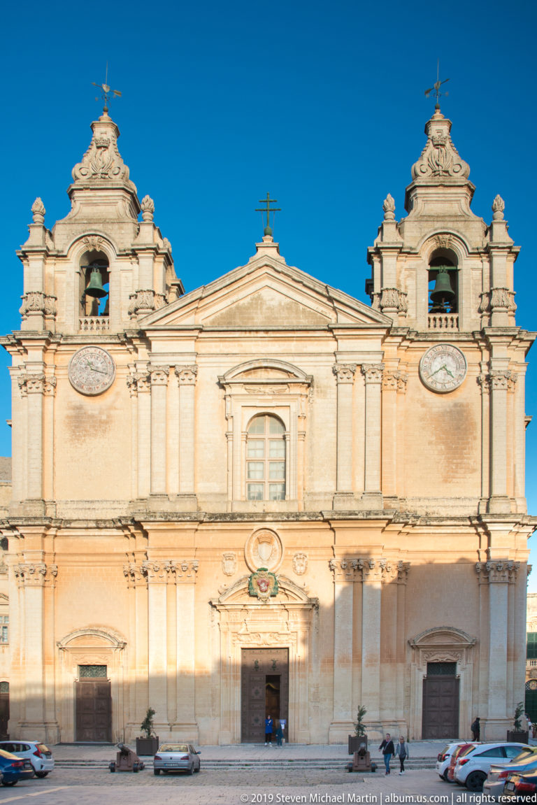 Full frontal view of St. Paul’s Cathedral with twin bell towers, clocks, central pediment, three entrances, and cars along the square.