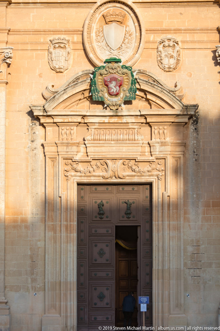 Central cathedral doorway with paneled doors, carved stone surround, colorful heraldic crest, and circular emblem above.
