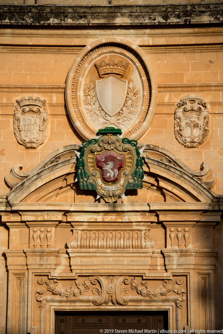 Close view of colorful crowned coat of arms beneath a circular stone emblem, flanked by smaller shields above a cathedral doorway.