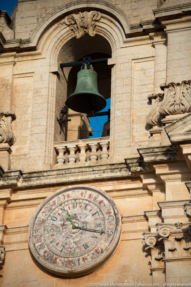 Large circular clock beneath an open belfry with bronze bell on the limestone tower of St. Paul’s Cathedral.