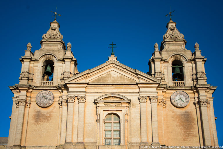 Upper façade of St. Paul’s Cathedral with twin bell towers, two clocks, central pediment, and pale limestone against blue sky.