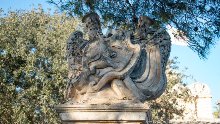 Stone heraldic sculpture with two winged figures supporting a large central shield on a pedestal beneath trees.