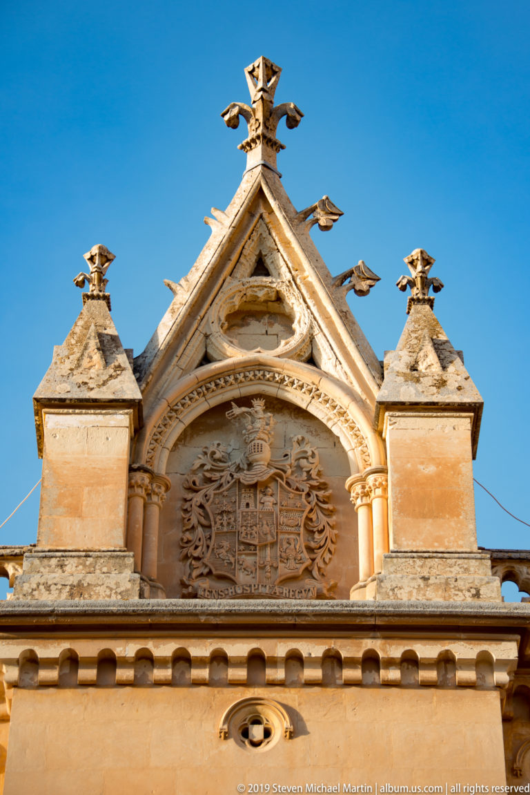 Ornate limestone gable with a circular carved heraldic relief, scrollwork, pinnacles, and decorative finials against blue sky.