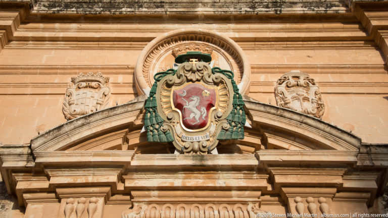 Colorful crowned coat of arms with green mantling above a stone pediment, flanked by smaller carved shields on a limestone façade.