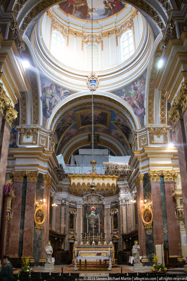 Church sanctuary viewed from the nave with colored-marble piers, dome, altar, hanging lamps, paintings, and statues.