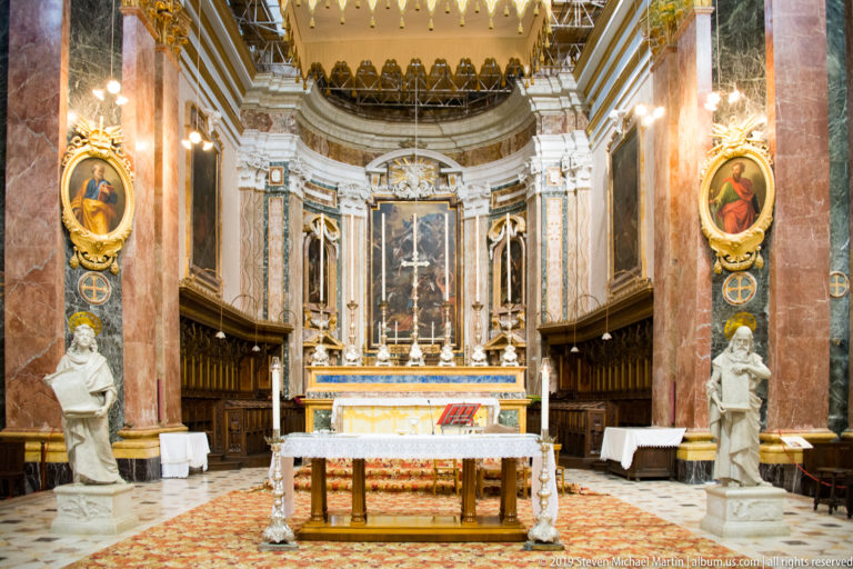 Cathedral sanctuary with marble altar, crucifix and altarpiece, dark choir stalls, white statues, oval paintings, and red-veined marble walls.