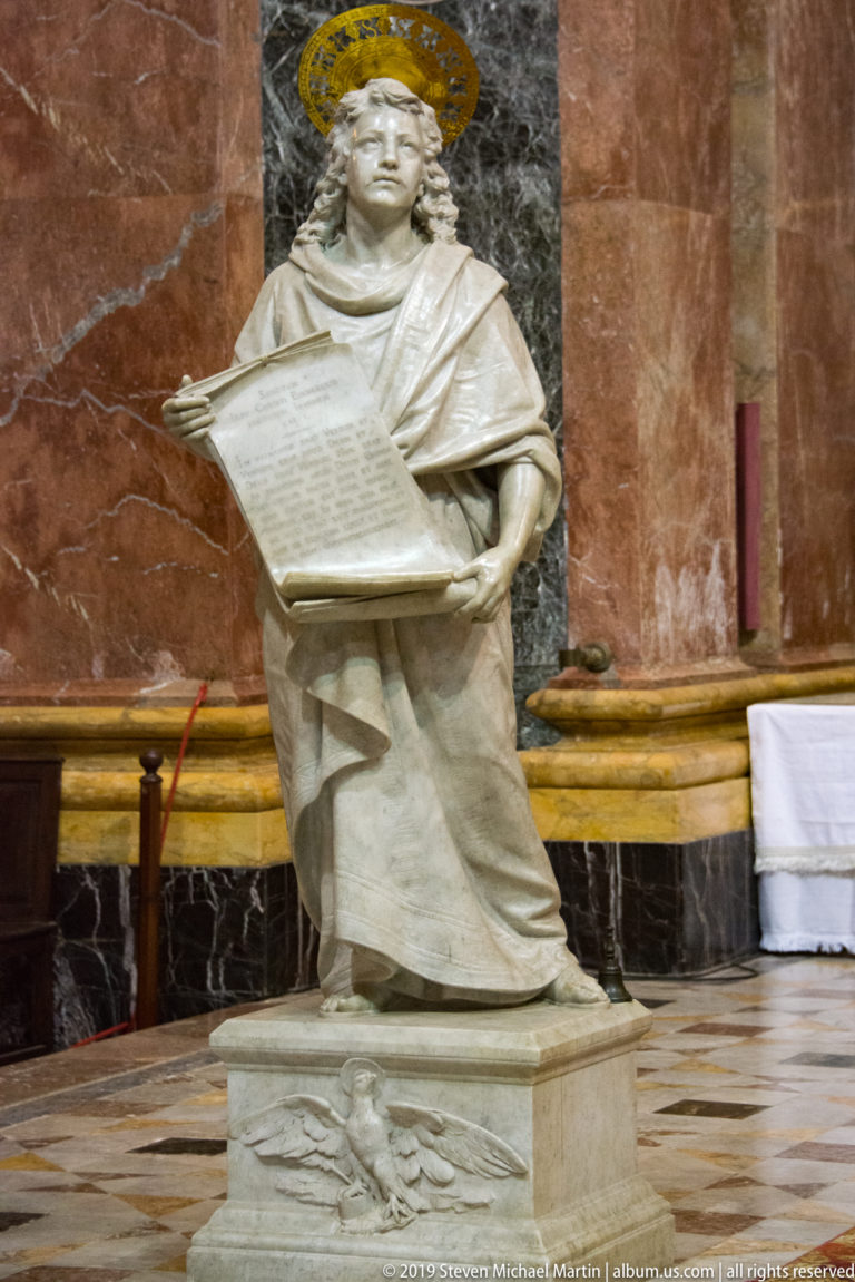Seated haloed marble figure holding an inscribed tablet against red and dark marble wall panels inside a church.