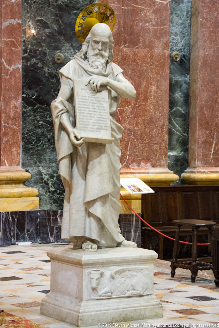 Haloed marble saint in clerical robes holding an inscribed tablet, standing on a carved pedestal against red and green marble.