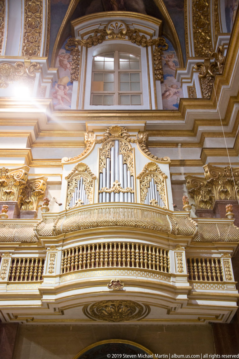 Pipe organ in a gilded case on an ornate gallery beneath a window, surrounded by painted and carved church decoration.