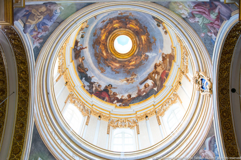 Close upward view of a frescoed dome centered on a bright oculus, with gilded ribs, painted figures, and drum windows.