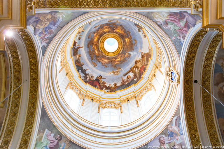 Upward view into a frescoed church dome with gilded rings, drum windows, painted figures, and a bright central oculus.
