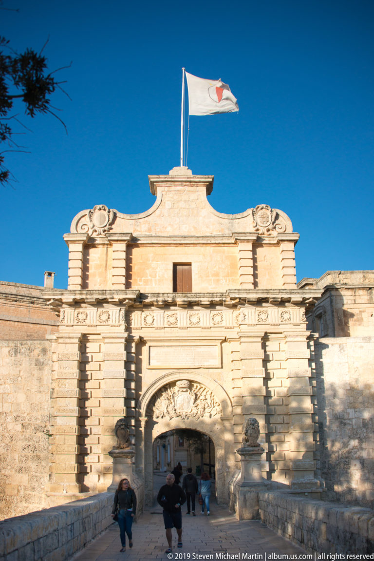 M’dina Gate with rusticated limestone façade, central arched passage, flag above, massive walls, bridge approach, and pedestrians entering.