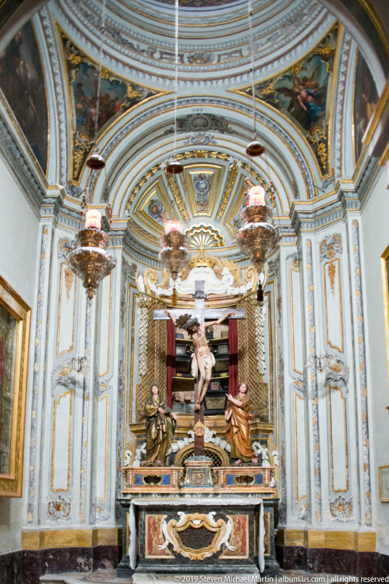 Side chapel with sculptural Crucifixion group above a marble altar, blue-gray paneled walls, gilded trim, paintings, and hanging lamps.