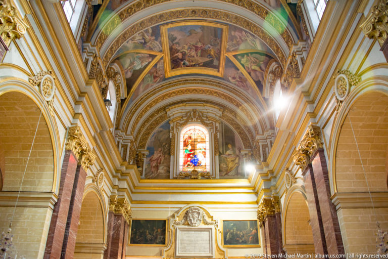 Ornate church nave with painted barrel vault, gilded moldings, side arches, framed paintings, and a stained-glass window.