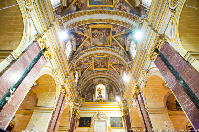 Upward view along a church nave with reddish marble piers, gilded painted vaults, arches, and a glowing stained-glass window.