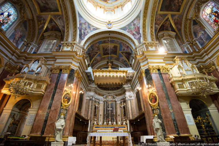 Symmetrical church sanctuary centered on a high altar, colored-marble columns, sculptures, painted vaults, and bright dome opening.