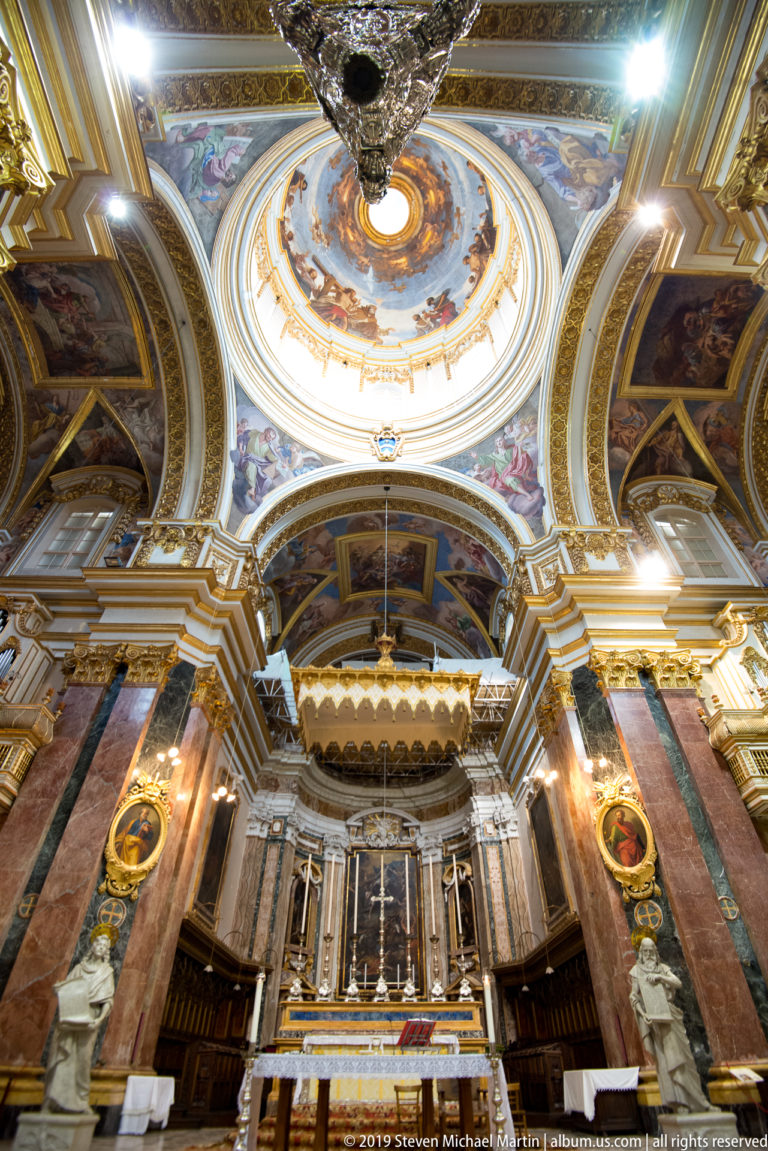Ornate church sanctuary with marble columns, sculpted figures, gilded details, monumental altar, and a painted dome overhead.