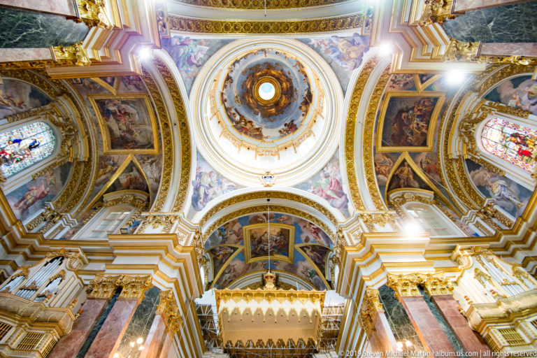 Wide upward church interior with central frescoed dome, painted vaults, stained glass, gilded moldings, marble piers, and organ loft.