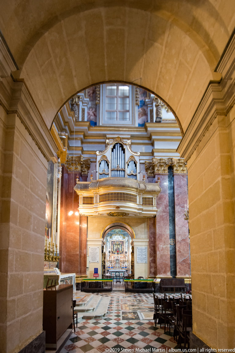 Wide limestone arch frames a richly decorated church interior with patterned floor, pews, organ loft, and illuminated altar.