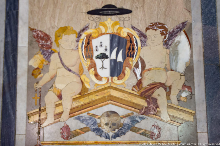 Painted heraldic shield flanked by two putti above a skull motif and crossed emblems in a framed wall panel.