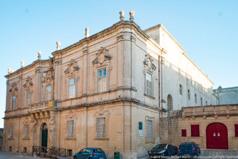 Large ornate limestone building with tall windows, sculpted surrounds, roof balustrade, and a long side elevation beside parked cars.