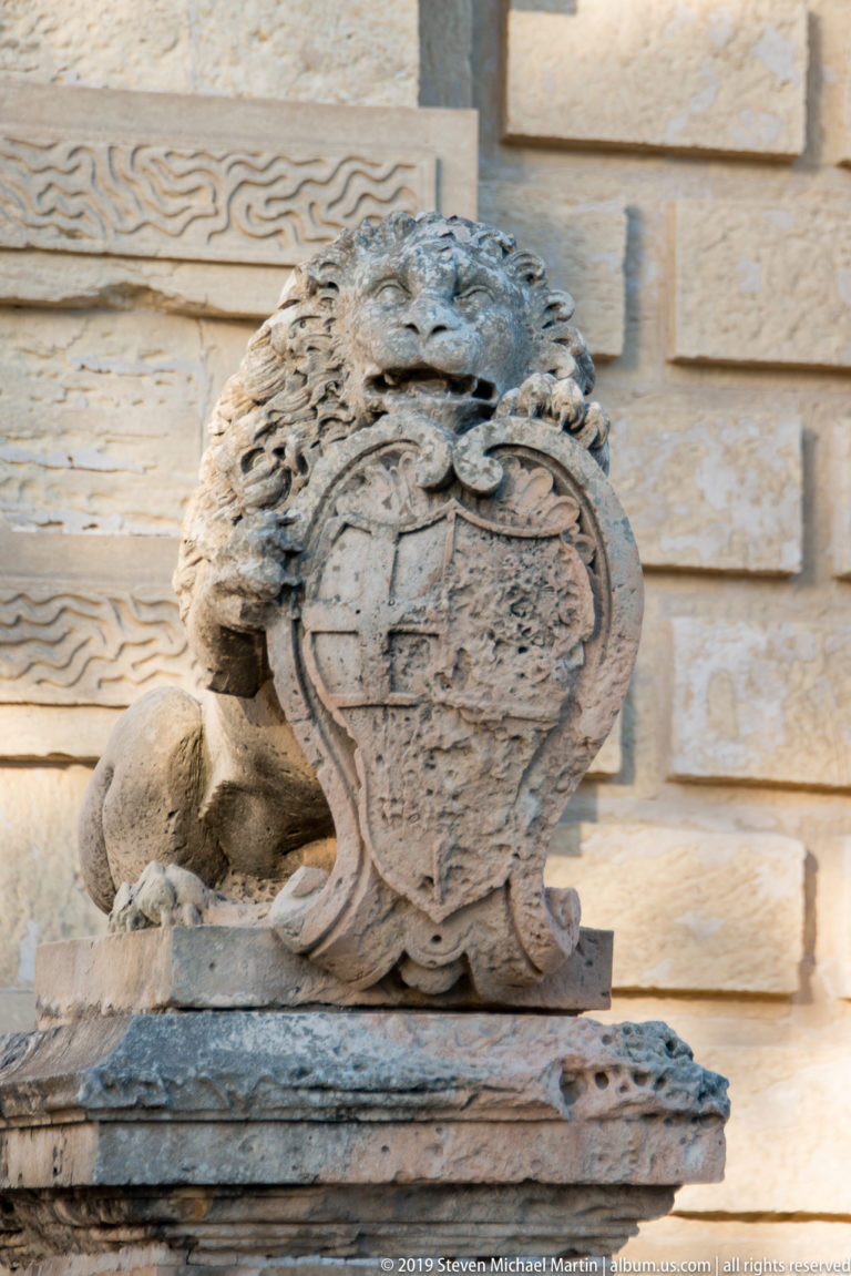 Close view of a weathered carved shield with curling ornament and a crouching stone supporter against rusticated limestone masonry.