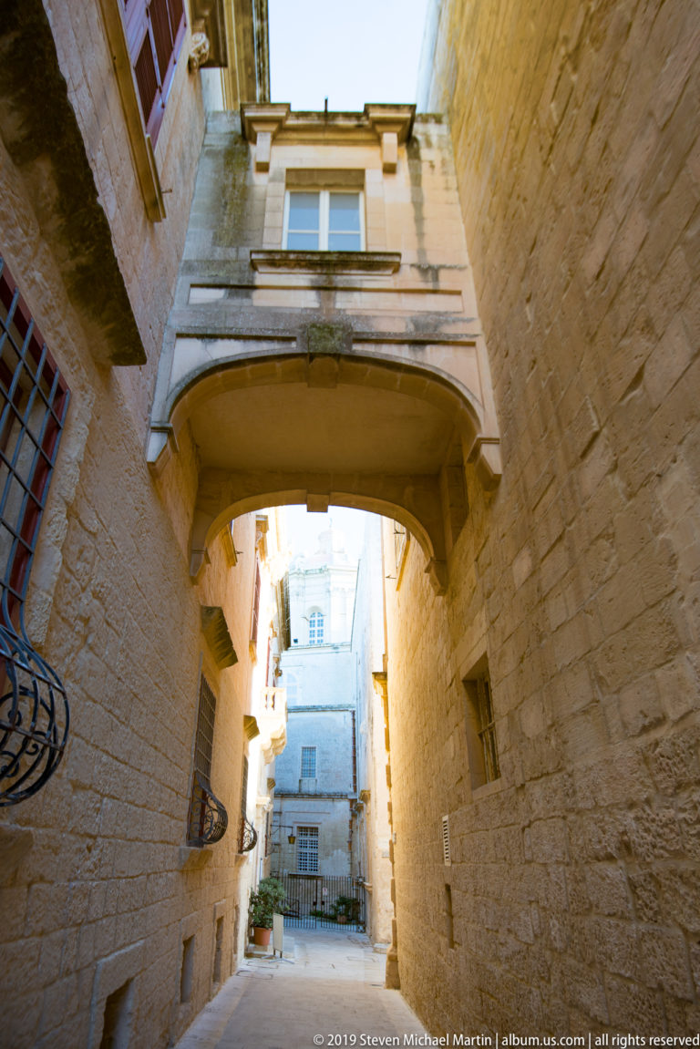 Covered bridge links limestone buildings above a narrow lane leading toward a bright courtyard with balconies and windows.