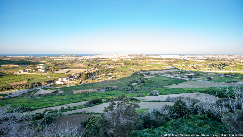Elevated view from M’dina across green fields, roads, scattered buildings, and distant Maltese settlements under a clear sky.
