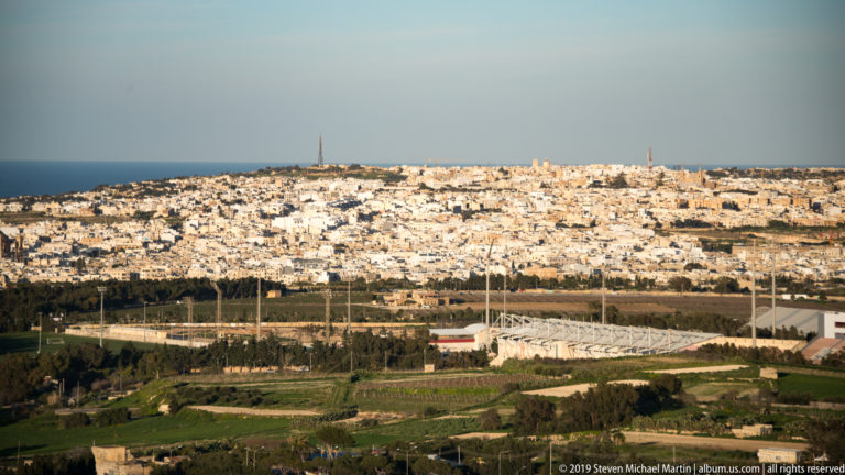 Elevated view across fields and marina masts toward a dense pale coastal settlement and the blue Mediterranean horizon.