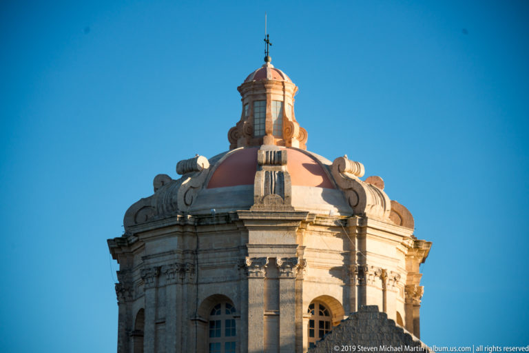 Ornate stone drum and reddish dome with columns, arched openings, scrolls, and a lantern against blue sky.