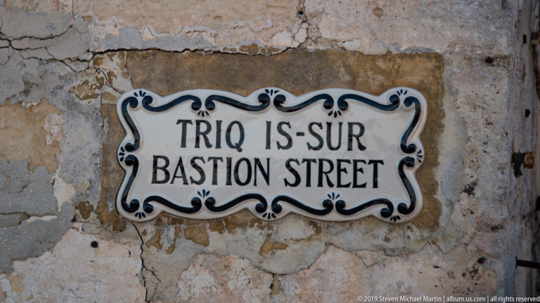 Blue-and-white ceramic plaque reading Triq Is-Sur, Bastion Street, mounted on weathered limestone.