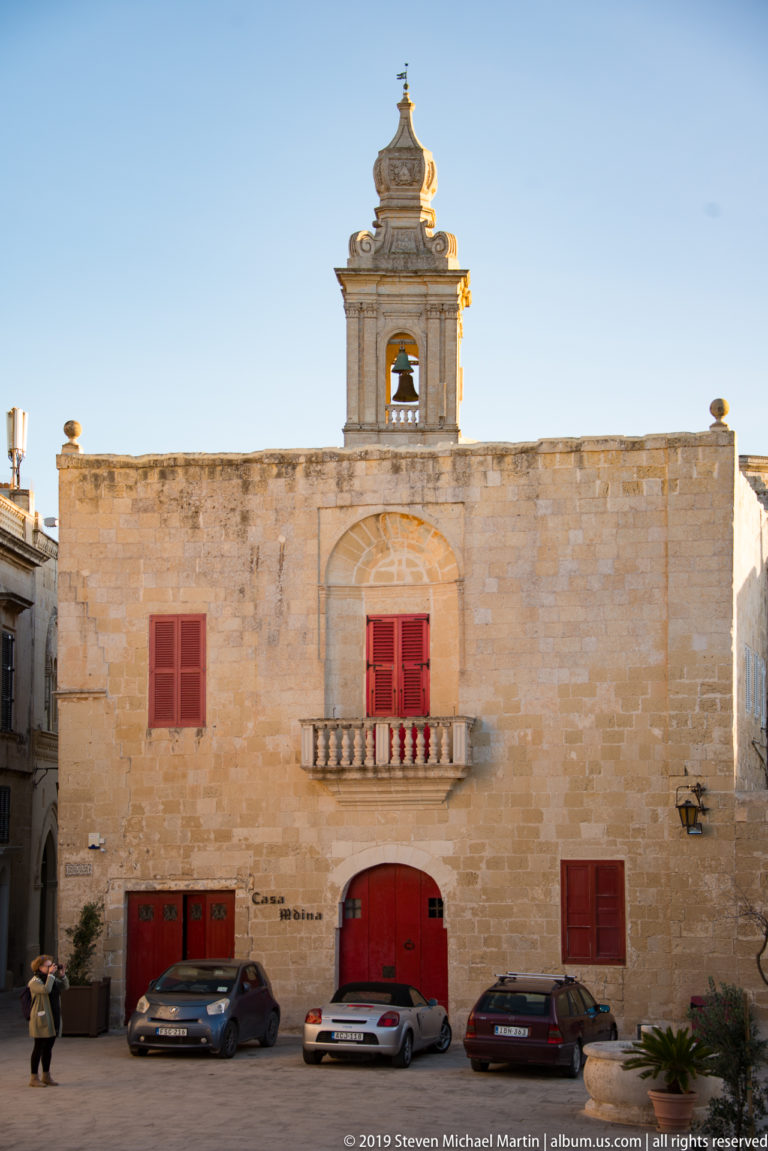 Symmetrical limestone façade with red shutters and doors, central balcony, and a small bell turret above an open square.
