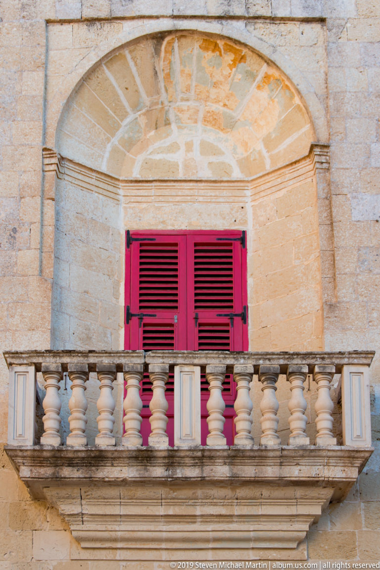 Stone balcony with turned balusters and bright red louvered shutters beneath a broad semicircular limestone arch.