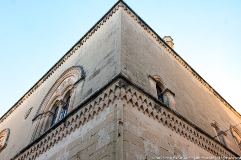 Upward view of a tall limestone building corner with arched window, pyramidal stone bossing, and projecting roofline.