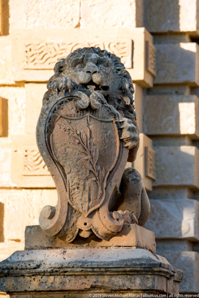 Weathered limestone heraldic shield surrounded by deeply carved leaves, scrolls, and shell-like ornament on an architectural pedestal.