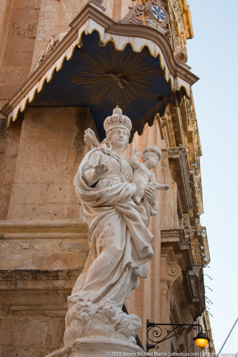 Crowned Madonna and Child stone statue beneath a dark canopy on the corner of an ornate limestone building.
