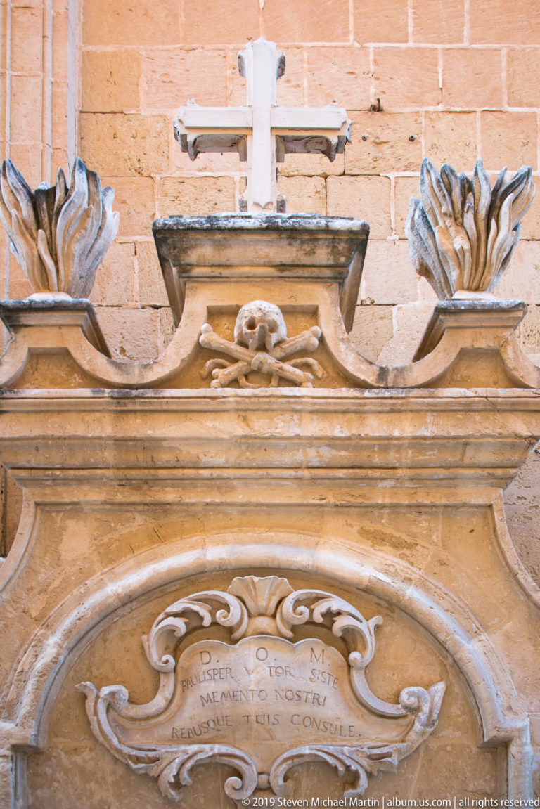 White cross above a carved skull, inscription panel, and two flaming urns on an ornate limestone architectural surround.